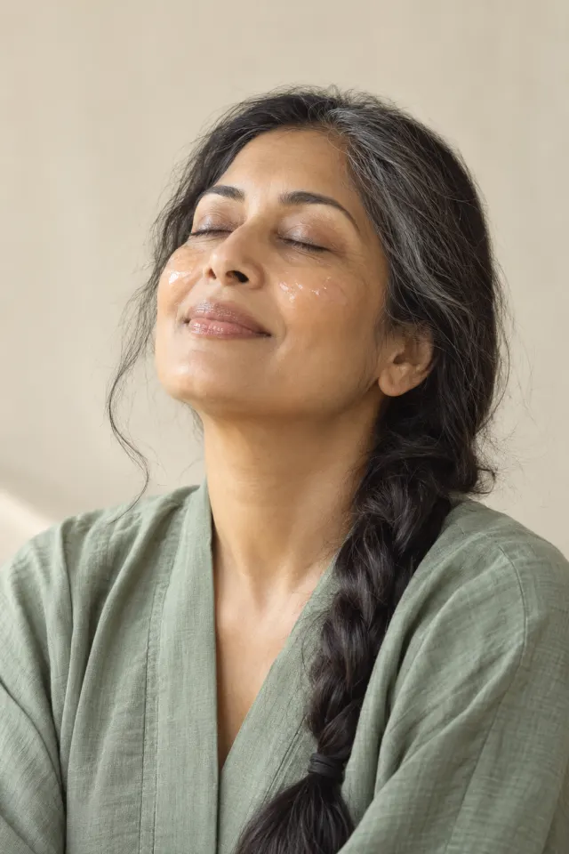 A woman with a long silver-streaked braid and closed eyes with a thin layer of clear gel on her cheekbones, in a sage robe.