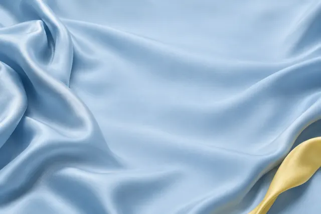 Studio photo of pale blue satin fabric in soft folds with a butter-yellow ribbon at the lower right.