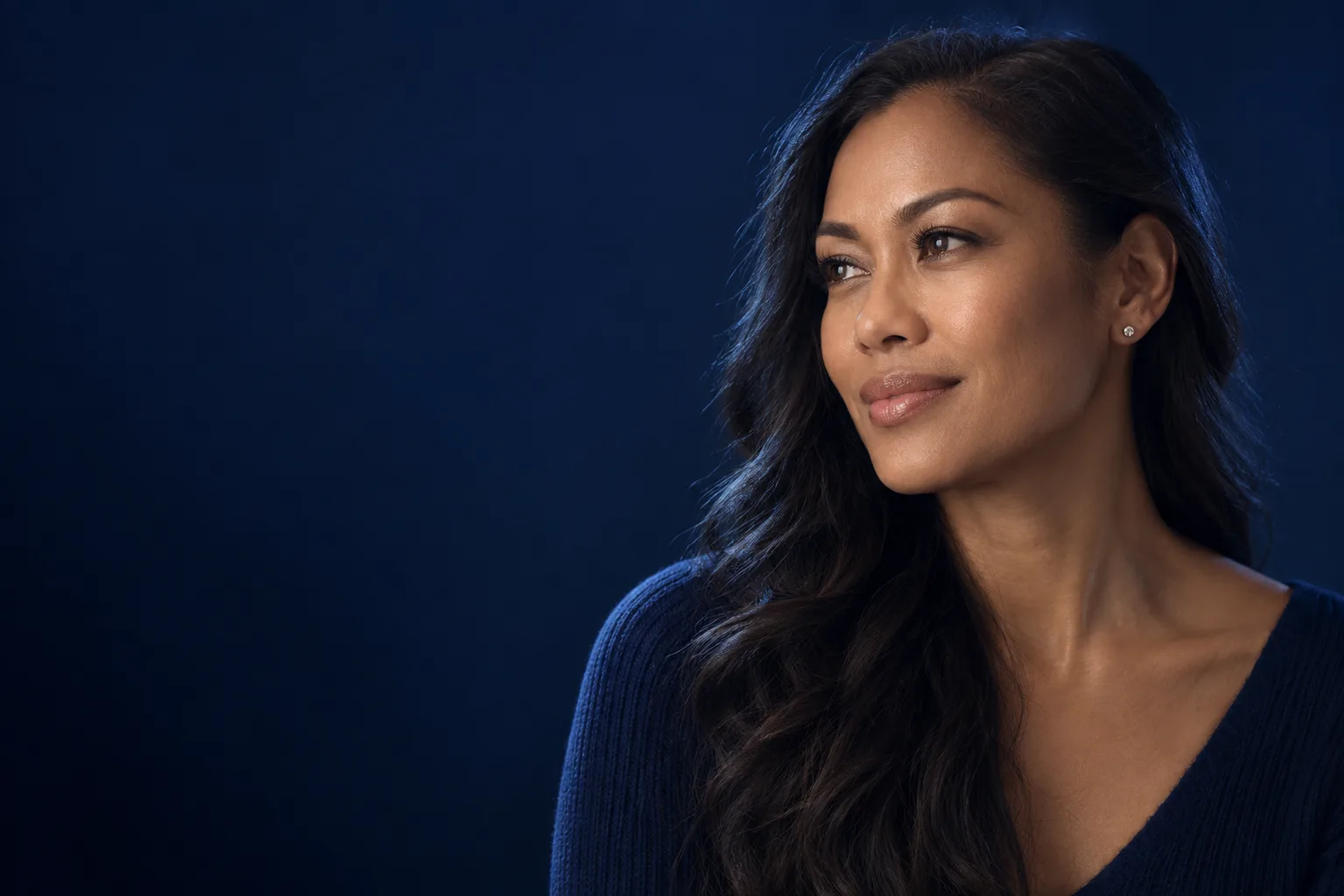 Editorial studio portrait of a woman with long dark wavy hair and a navy ribbed top, looking away to the left with a calm half-smile against a deep navy backdrop.