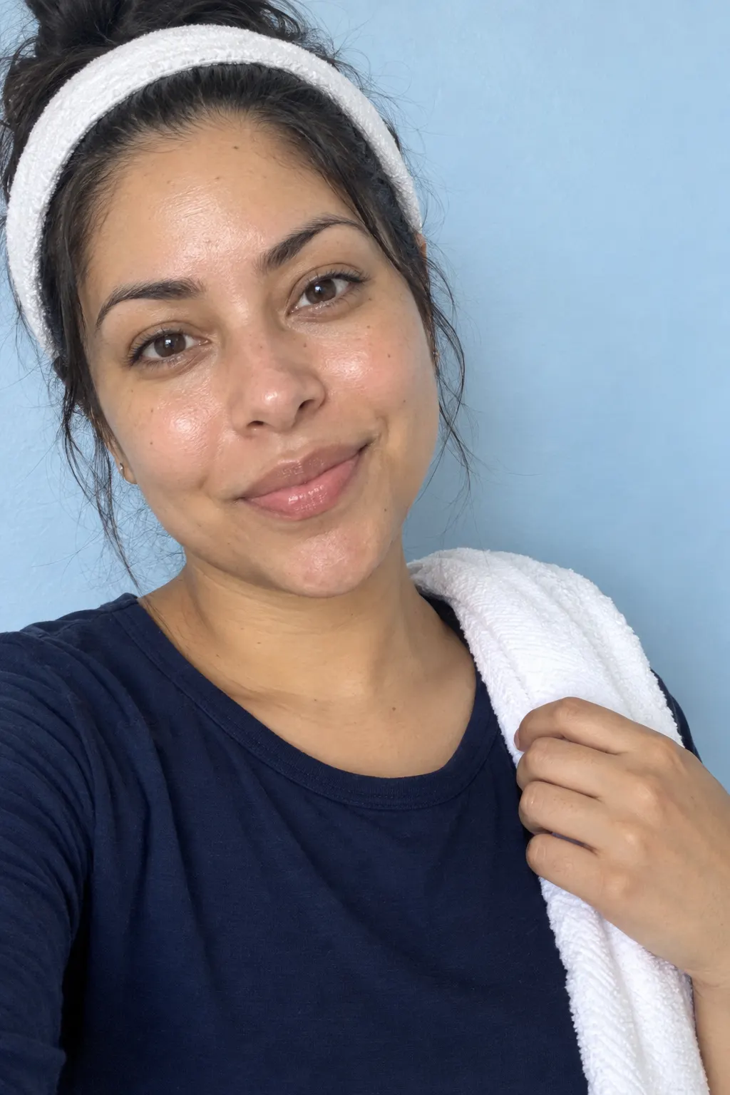 Casual snapshot of a smiling woman with a white headband and a towel on her shoulder against a pale blue wall.